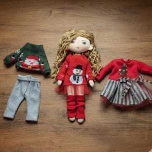 Peut inclure: Une poup&eacute;e faite &agrave; la main avec des cheveux blonds boucl&eacute;s, v&ecirc;tue d'un pull rouge avec un bonhomme de neige, de leggings rouges et d'une jupe en tulle rouge. D'autres tenues incluent un pull vert du P&egrave;re No&euml;l et un pantalon gris, et une robe rouge avec une jupe ray&eacute;e.