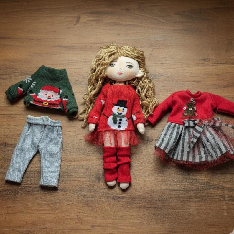 Peut inclure: Une poup&eacute;e faite &agrave; la main avec des cheveux blonds boucl&eacute;s, v&ecirc;tue d'un pull rouge avec un bonhomme de neige, de leggings rouges et d'une jupe en tulle rouge. D'autres tenues incluent un pull vert du P&egrave;re No&euml;l et un pantalon gris, et une robe rouge avec une jupe ray&eacute;e.