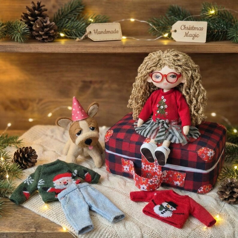 Peut inclure: Une poup&eacute;e faite &agrave; la main avec des cheveux blonds boucl&eacute;s et des lunettes rouges, portant un pull rouge avec un motif de sapin de No&euml;l vert et une jupe &agrave; rayures grises et blanches. Un chien en peluche avec un chapeau de f&ecirc;te et des pulls et pantalons de No&euml;l miniatures sont &eacute;galement pr&eacute;sents. La poup&eacute;e est assise sur une valise &agrave; carreaux rouge et noir.
