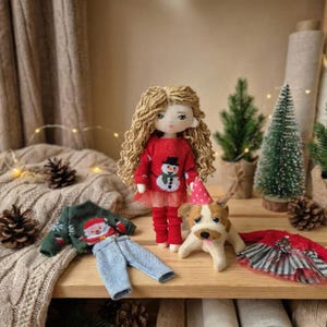 Peut inclure: Une poup&eacute;e faite &agrave; la main avec de longs cheveux blonds boucl&eacute;s portant un pull rouge avec un motif de bonhomme de neige, des leggings rouges et un chapeau de f&ecirc;te rose. Des v&ecirc;tements de poup&eacute;e suppl&eacute;mentaires et une figurine de chien sont &eacute;galement pr&eacute;sents, ainsi que des sapins de No&euml;l miniatures.