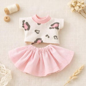 May include: A doll-sized outfit featuring a white t-shirt with pink and gray leopard print and a pink skirt. The t-shirt has a pink collar and short sleeves. The skirt is a gathered style. The outfit is designed for a doll.
