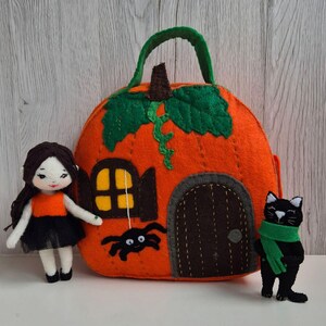 Op de afbeelding: Een vilten Halloween-set met een oranje pompoenvormig huis met een groene handgreep en bladeren. Een pop in een oranje en zwarte outfit en een zwarte kat met een groene sjaal staan naast het huis. Een zwarte spin hangt uit het raam.