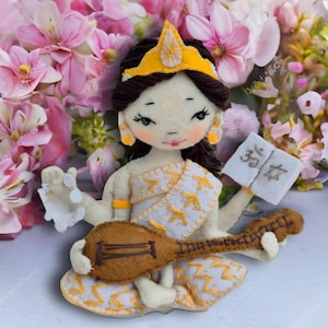 May include: A felt doll with a white and yellow dress, a gold crown, and a brown musical instrument. The doll is holding a white sign with the text "35" on it. The doll is surrounded by pink flowers.