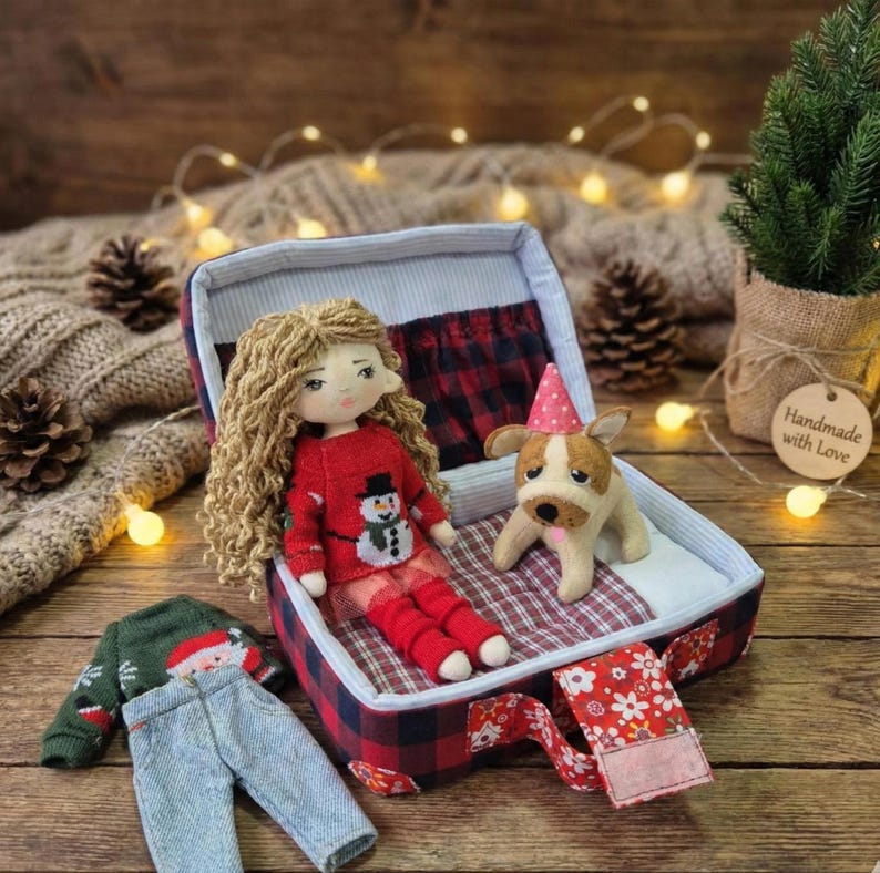 Peut inclure: Une poup&eacute;e faite &agrave; la main avec de longs cheveux blonds boucl&eacute;s, v&ecirc;tue d'un pull rouge avec un motif de bonhomme de neige et un pantalon rouge, assise dans une valise &agrave; carreaux rouges et noirs. Un petit chien avec un chapeau de f&ecirc;te et une tenue miniature sont &eacute;galement pr&eacute;sents.