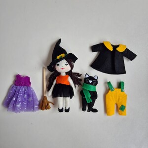 Op de afbeelding: Een verzameling handgemaakte Halloween-thema vilten poppen en accessoires. Inclusief een pop verkleed als heks met een zwarte hoed, een zwarte kat met een groene sjaal, een paarse jurk, een bezem, een zwarte jas en gele tuinbroek.