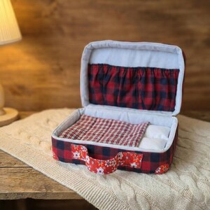 Peut inclure: Un petit lit pour animaux en forme de valise, avec un motif &agrave; carreaux rouges et noirs. L'int&eacute;rieur comprend une couverture &agrave; carreaux rouges et blancs et un petit oreiller blanc. La poign&eacute;e est orn&eacute;e de fleurs.