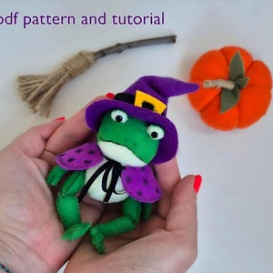 May include: A handmade Halloween-themed felt frog wearing a purple witch hat and cape with polka dots. An orange felt pumpkin and a small broom are also in the image. The text "pdf pattern and tutorial" is at the top.