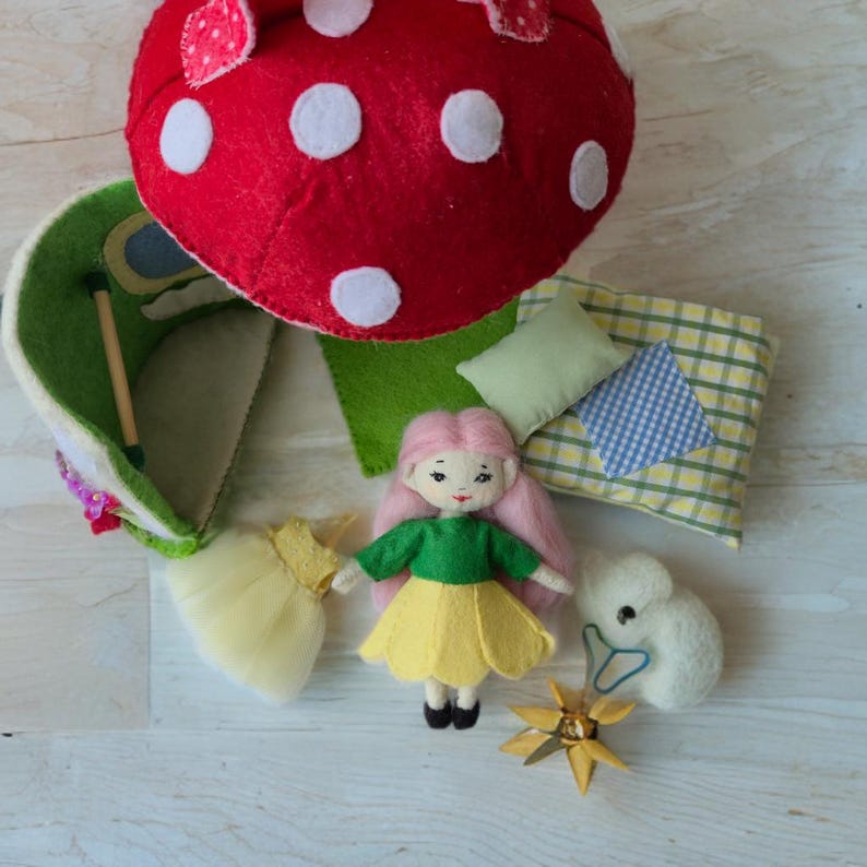 May include: A handmade felt doll with pink hair, a green top, and a yellow skirt stands near a red mushroom house with white spots. Other felt accessories include a yellow dress, a white bunny, pillows, and a gold star. The scene is set on a light wood surface.
