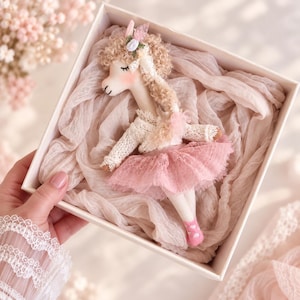 Peut inclure: Une poupée lama en tissu faite à la main avec des cheveux beiges bouclés, portant un haut en dentelle blanche et une jupe en tulle rose à volants. La poupée a des yeux fermés brodés, des joues roses et des chaussettes roses à cœurs.
