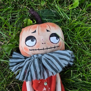 Puede incluir: Una mu&ntilde;eca de calabaza hecha a mano con una cara cosida, ojos grandes y una boca cosida. La mu&ntilde;eca tiene un cuello a rayas y un vestido rojo con un dise&ntilde;o de copo de nieve blanco. La mu&ntilde;eca est&aacute; tumbada en hierba verde.