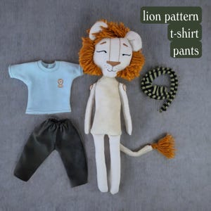 May include: A plush lion doll with a light brown body, a fluffy orange mane, and a long tail. The doll is wearing a light blue t-shirt with a small lion graphic, dark green pants, and a striped scarf. The text "lion pattern t-shirt pants" is written in black on a white background.