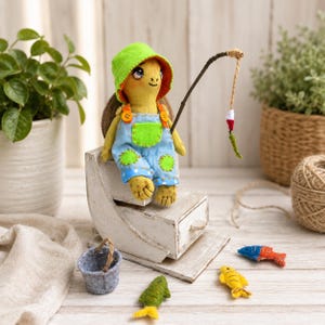 May include: A fabric turtle doll wearing a bright green hat and blue overalls with orange buttons and green patches. The doll holds a thin wooden fishing rod with a string and a small float. Nearby are three small felt fish and a grey felt bucket.