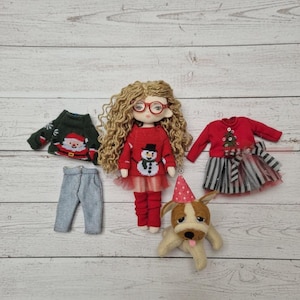Peut inclure: Une poup&eacute;e aux cheveux blonds boucl&eacute;s et aux lunettes rouges, v&ecirc;tue d'un pull rouge avec un bonhomme de neige. Des v&ecirc;tements de poup&eacute;e suppl&eacute;mentaires comprennent un pull vert du P&egrave;re No&euml;l, un pantalon gris et une robe rouge. Un petit chien en peluche avec un chapeau de f&ecirc;te est &eacute;galement pr&eacute;sent.