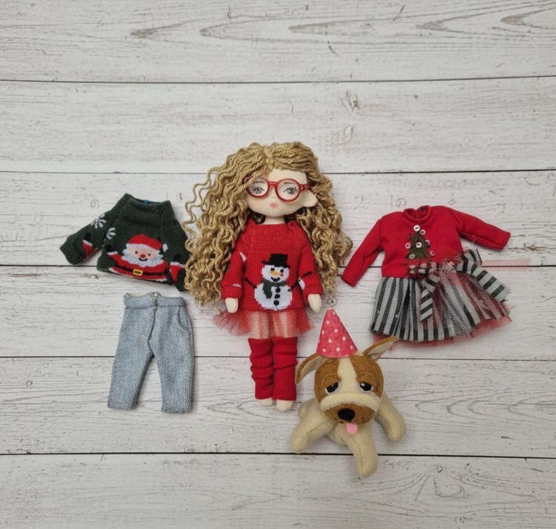 Peut inclure: Une poup&eacute;e aux cheveux blonds boucl&eacute;s et aux lunettes rouges, v&ecirc;tue d'un pull rouge avec un bonhomme de neige. Des v&ecirc;tements de poup&eacute;e suppl&eacute;mentaires comprennent un pull vert du P&egrave;re No&euml;l, un pantalon gris et une robe rouge. Un petit chien en peluche avec un chapeau de f&ecirc;te est &eacute;galement pr&eacute;sent.