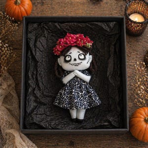 May include: A handmade doll with a Day of the Dead theme, featuring a floral crown and a patterned dress. The doll is presented in a black box with a textured background, alongside pumpkins and a lit candle.