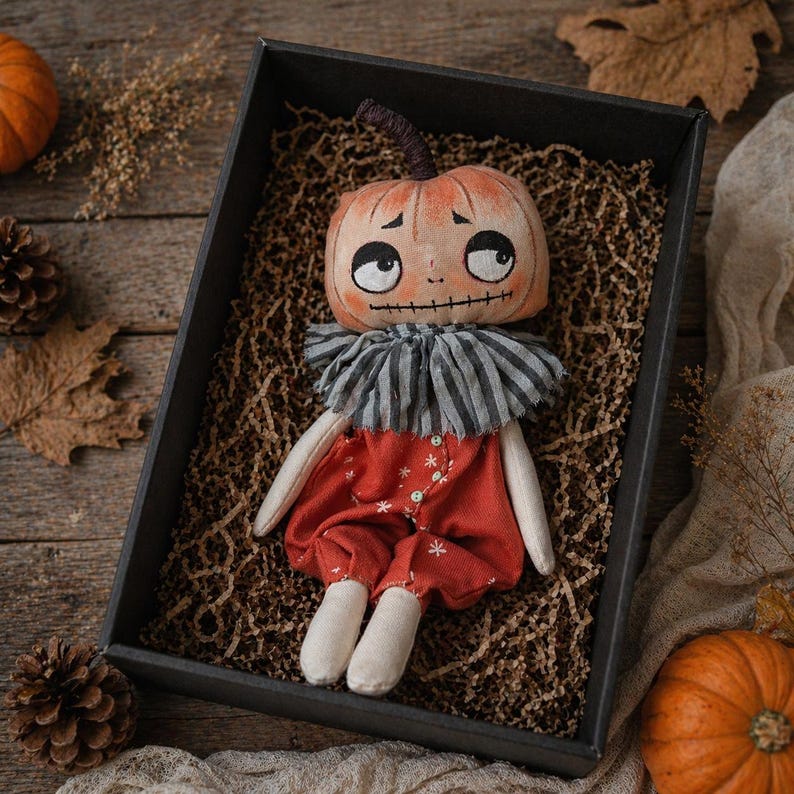 Puede incluir: Mu&ntilde;eca de calabaza hecha a mano con una cara pintada, vestida con un atuendo rojo con detalles de copos de nieve blancos y un cuello a rayas. La mu&ntilde;eca se presenta en una caja negra con relleno de papel marr&oacute;n, evocando un tema de oto&ntilde;o o Halloween.