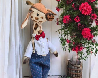 May include: A plush giraffe toy wearing a white shirt, blue pants with white polka dots, and a red bow tie. The giraffe is standing in front of a white door and a potted plant with red flowers.