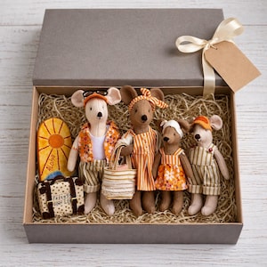 Puede incluir: Una caja de regalo gris con cuatro muñecos de ratón vestidos con ropa de vacaciones. Los ratones llevan gafas de sol, sombreros y trajes en naranja, marrón y verde. Los accesorios incluyen una tabla de surf, una maleta y un bolso de mano. La caja está atada con una cinta y una etiqueta en blanco.