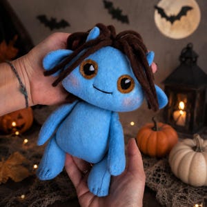 Puede incluir: Una muñeca de peluche azul con cabello de lana marrón oscuro, ojos grandes de color marrón y orejas puntiagudas. La muñeca tiene una textura suave similar al fieltro, una boca pequeña cosida y una forma corporal humanoide simple.