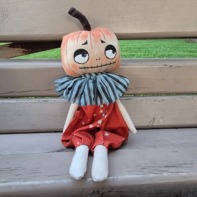 Puede incluir: Mu&ntilde;eca hecha a mano con cabeza en forma de calabaza y expresi&oacute;n triste. La mu&ntilde;eca tiene cuello a rayas, peto rojo con detalles de flores blancas y calcetines blancos. La mu&ntilde;eca est&aacute; sentada en un banco de madera.