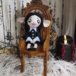 May include: A plush doll with pale skin, large dark eyes, and black braided hair, wearing a black dress with a white collar and cuffs. The doll has prominent ears and sits on a small, ornate wooden chair with a high back and curved armrests.