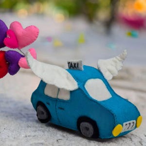 May include: A blue felt toy car with white wings and the word "TAXI" on top. The car is being pulled by a string of felt hearts in pink, red, and purple.