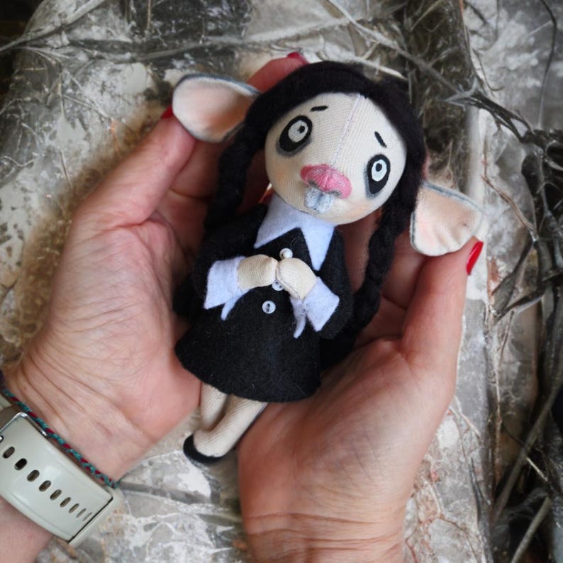 May include: A handmade fabric doll featuring a pale face with large dark eyes, a pink nose, and two prominent front teeth. The doll has black hair styled in two long braids and wears a black dress with a white collar and white cuffs with buttons.