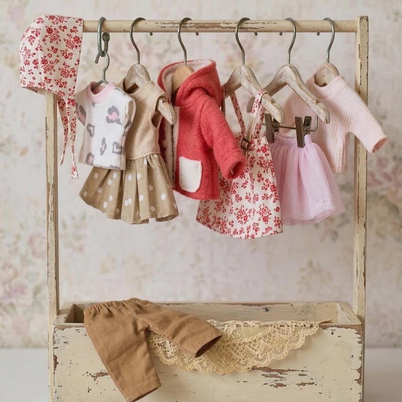 May include: A miniature clothing set for a doll, including a bonnet, a top, a skirt, a jacket, a dress, a skirt, a sweater, and pants. The clothes are in various colors, including pink, beige, and red, and are hanging on a small wooden rack.