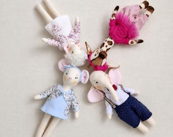 May include: Four plush animal dolls arranged in a circle. One is a white sheep in a floral dress, one is a giraffe in a pink top and striped pants, one is a mouse in a blue floral dress, and one is an elephant in a white shirt and blue pants.
