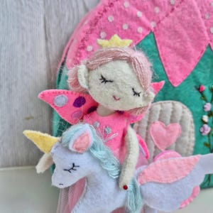 Pu&ograve; includere: Una bambola di fata in feltro fatta a mano con ali rosa e una corona d'oro cavalca un unicorno bianco con criniera blu e ali rosa. La fata indossa un vestito rosa. Sullo sfondo c'&egrave; una casa di feltro rosa e verde.