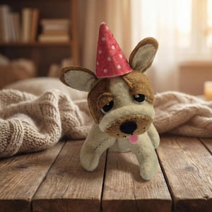 Peut inclure: Un chien en peluche fait &agrave; la main, avec une fourrure marron et blanche, portant un chapeau de f&ecirc;te rouge &agrave; pois blancs. Le chien a une expression triste et une langue rose. Il est pos&eacute; sur une surface en bois.
