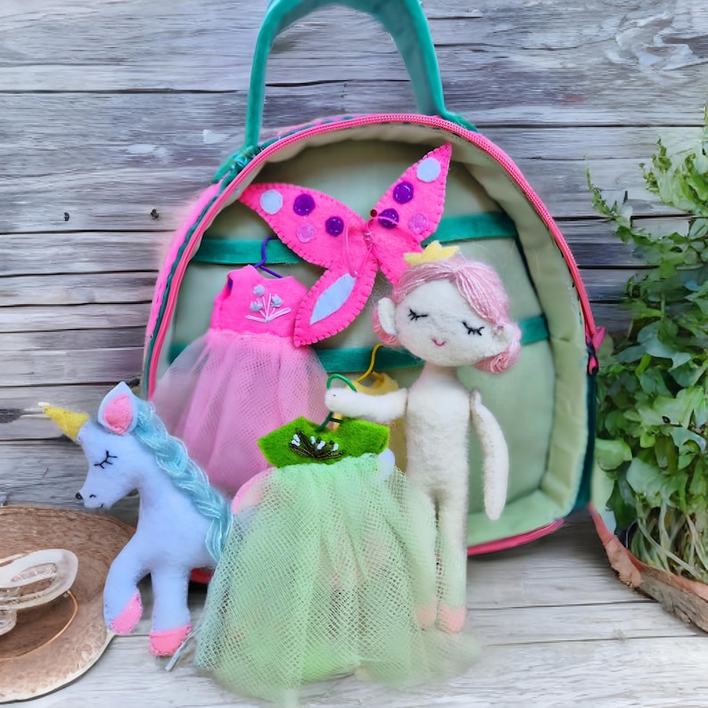 Pu&ograve; includere: Una bambola di feltro con capelli rosa e una corona, che tiene in mano un vestito verde. Un unicorno blu, abiti rosa e una decorazione a farfalla sono vicino a una borsa verde e rosa. La scena evoca un tema da favola.