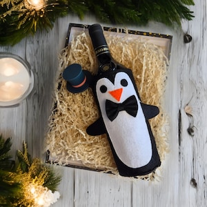 May include: A wine bottle wrapped in a black and white penguin-shaped felt cover with a black bow tie and orange beak. The bottle is in a brown paper-filled gift box with a small black top hat.