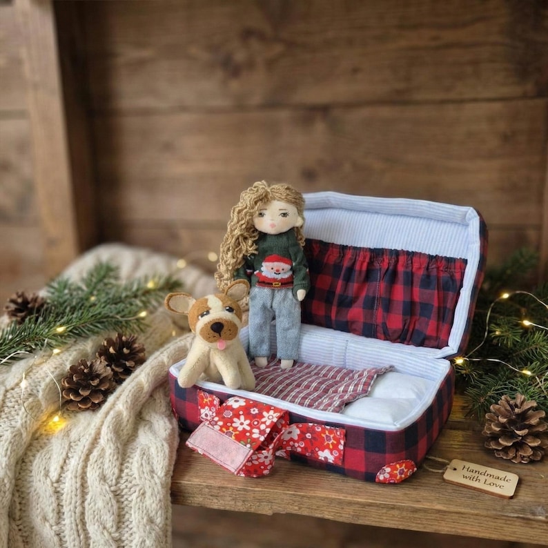 Peut inclure: Une poup&eacute;e faite &agrave; la main avec de longs cheveux boucl&eacute;s, portant un pull vert avec un motif de P&egrave;re No&euml;l et un pantalon gris, se tient dans une valise ouverte. Un chien en peluche est assis &agrave; c&ocirc;t&eacute; de la poup&eacute;e. La valise est &agrave; carreaux rouges et noirs.