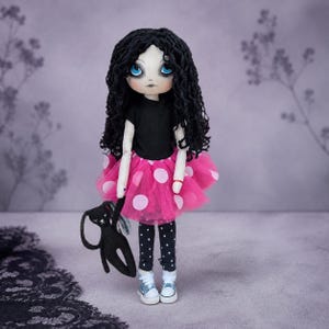 Pode incluir: Uma boneca de pano feita à mão com longos cabelos pretos cacheados e grandes olhos azuis. A boneca veste uma camiseta preta de manga curta, uma saia de tule rosa vibrante com bolinhas brancas, leggings pretas com bolinhas brancas e tênis azuis.