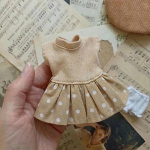 May include: A miniature doll dress with a beige bodice and a pleated skirt with white polka dots. The dress has a high neckline and is paired with white underwear. The items are on a background of vintage sheet music.
