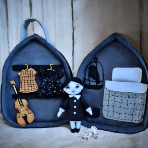Op de afbeelding: Een handgemaakte pop met zwart haar en een zwarte jurk staat voor een open, donkerblauwe vilten draagtas. De tas heeft een miniatuur garderobe met twee jurken, een viool, een vogelkooi en een bed. Een klein wit speeltje ligt aan de voeten van de pop.