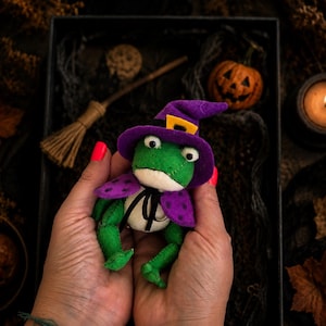 May include: A small green felt frog figurine wearing a purple witch hat with a yellow buckle and a matching purple cape with black polka dots. The frog has a white belly, black stitched mouth, and black ribbon ties securing the cape around its neck.