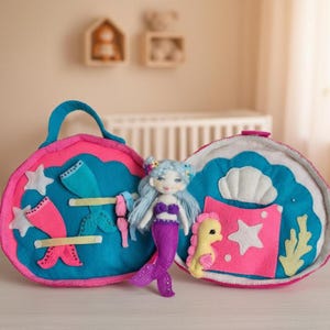 Può includere: Una bambola sirena in feltro con capelli blu e coda viola è seduta all'interno di una valigetta in feltro rosa e blu aperta. La custodia presenta stelle e conchiglie, e un cavalluccio marino giallo.