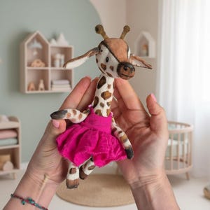 Puede incluir: Una muñeca jirafa hecha a mano con un patrón de manchas marrones y blancas, que lleva un tutú rosa brillante. La jirafa tiene orejas marrones y un hocico marrón. La muñeca se sostiene frente a un fondo suave verde y blanco, lo que sugiere una habitación infantil.