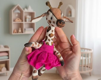 May include: A handmade giraffe doll with a brown and white spotted pattern, wearing a bright pink tutu. The giraffe has brown ears and a brown snout. The doll is held in front of a soft green and white background, suggesting a nursery or children's room.