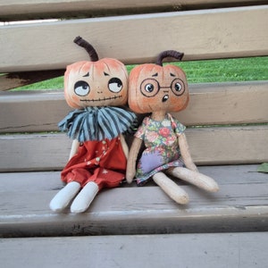 Puede incluir: Dos mu&ntilde;ecas hechas a mano con cabezas de calabaza, caras pintadas y ropa de tela. Una mu&ntilde;eca viste un atuendo rojo con detalles blancos y un cuello a rayas azul y blanco. La otra mu&ntilde;eca viste un vestido con estampado floral y gafas.