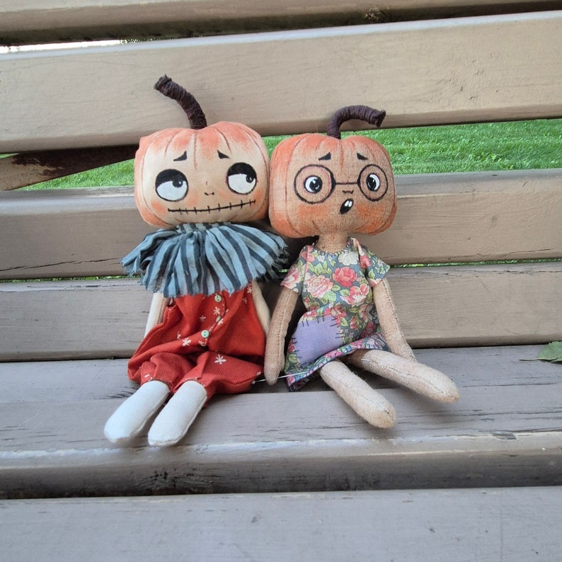 Puede incluir: Dos mu&ntilde;ecas hechas a mano con cabezas de calabaza, caras pintadas y ropa de tela. Una mu&ntilde;eca viste un atuendo rojo con detalles blancos y un cuello a rayas azul y blanco. La otra mu&ntilde;eca viste un vestido con estampado floral y gafas.