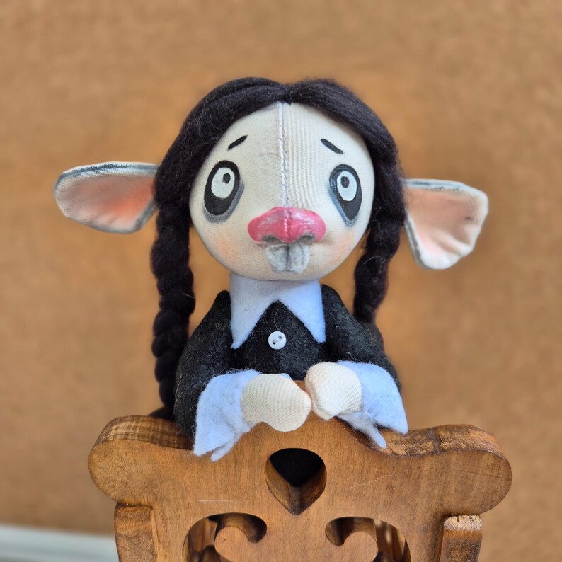 May include: A handmade fabric doll with a pale face, large dark eyes, a pink nose, and two black yarn braids. The doll wears a black dress with a white collar and cuffs, featuring a single white button on the front. The doll is positioned behind a carved wooden chair back.