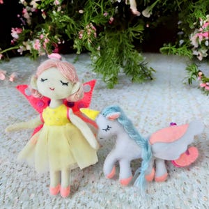 Pu&ograve; includere: Una bambola fata di feltro con capelli rosa, un vestito giallo e ali rosa &egrave; in piedi accanto a un unicorno bianco con una criniera blu e zoccoli rosa. La fata ha gli occhi chiusi e sorride. Anche l'unicorno ha gli occhi chiusi.