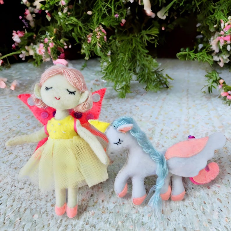 Pu&ograve; includere: Una bambola fata di feltro con capelli rosa, un vestito giallo e ali rosa &egrave; in piedi accanto a un unicorno bianco con una criniera blu e zoccoli rosa. La fata ha gli occhi chiusi e sorride. Anche l'unicorno ha gli occhi chiusi.