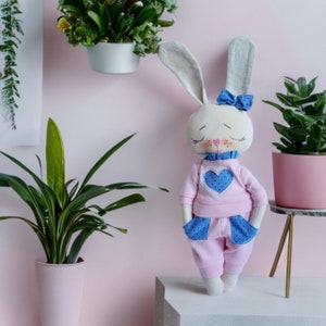 May include: A handmade plush bunny toy wearing a pink and blue outfit with a heart design. The bunny has a white face with closed eyes and a blue bow.