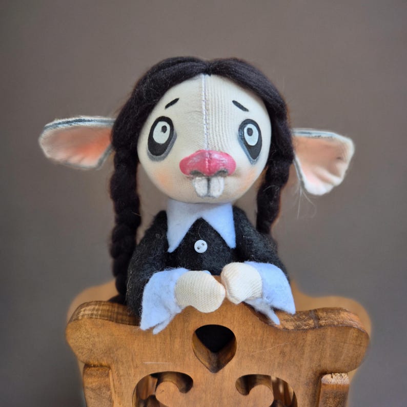 May include: A handmade fabric doll with a pale face, large dark eyes, a pink nose, and two front teeth. The doll has black yarn hair styled in two braids and wears a black long-sleeved garment with a white collar and white cuffs on the sleeves.
