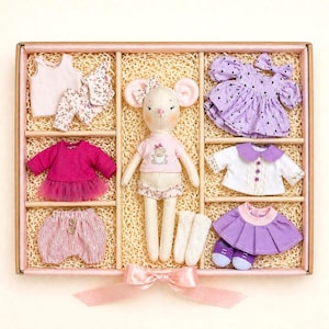 May include: A doll with a mouse face and several outfits in a box. The outfits include a pink top and shorts, a purple dress with black polka dots, a white jacket with a purple collar, and a pink skirt. The box is tied with a pink ribbon.