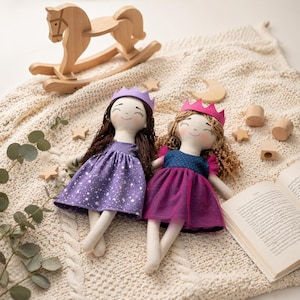 Può includere: Due bambole principessa fatte a mano con corone e vestiti, una viola e una rosa, giacciono su una coperta color crema. Un cavallo a dondolo di legno e altri giocattoli di legno sono sullo sfondo. Un libro per bambini è aperto nelle vicinanze.